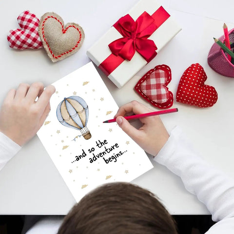 Child's hands holding Hot Air Balloon Adventure Baby Shower Card For New Parents Funny Congratulations Card Blank Inside 8x5.3 Inch Size With Envelope Perfect For Pregnant Couples And Newborn Celebrations surrounded by hearts and gifts.