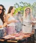 Group of friends enjoying a barbecue outdoors on a sunny day with grilled meats and casual summer attire.
