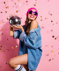 Person holding disco ball with star confetti, wearing denim and pink accessories, celebrating with joy against pink backdrop.