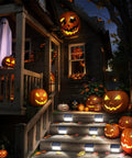 Halloween-decorated porch and steps with pumpkins and ghosts, illuminated by Outdoor Solar Deck Lights 6 Pack Waterproof Fence Post Lights For Backyard Patio Stair Wall Lighting With 3 LED Cool White Beams Solar Powered Panels For Quick Charging And No Fuss Install Up To 8 Hours Of Bright Illumination.