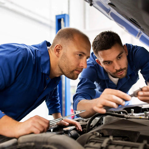 Two mechanics in blue uniforms using tools under a car hood, focusing on repairs.