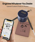 Front view of LaserPecker LP1 Pro laser engraver, blue top on tripod, engraving brown notebook; smartphone shows design app.