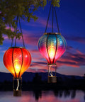 Set Of 2 Dope Hot Air Balloon Solar Lanterns For Outdoor Garden Decor, glowing in sunset, hanging from tree branches by water.