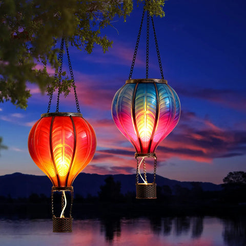 Set Of 2 Dope Hot Air Balloon Solar Lanterns For Outdoor Garden Decor, glowing in sunset, hanging from tree branches by water.