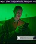 A person using a smartphone with a smart home security app interface, green ambient lighting in the room.