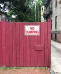 Heavy Duty Aluminum No Dumping Signs Two Pack on a red wooden fence in an urban area, preventing illegal dumping.