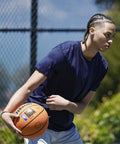 A person playing basketball outdoors with the Official DRV Series Basketball 27.5 Inch Size 5 In Brown With Rugged Outdoor Cover And NBA Branding.