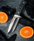 Survival Knife For Camping And Bushcraft With 14.5 cm Drop Point Blade In Stainless MOVA 1.4116 next to orange slices and sheath