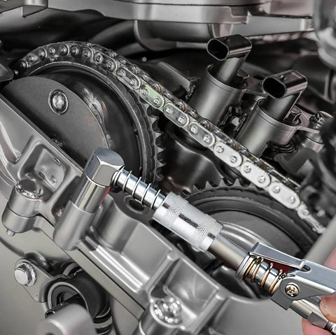 Close-up of a silver 90-degree grease gun adapter connected to a grease gun beside a motorcycle chain and sprocket.