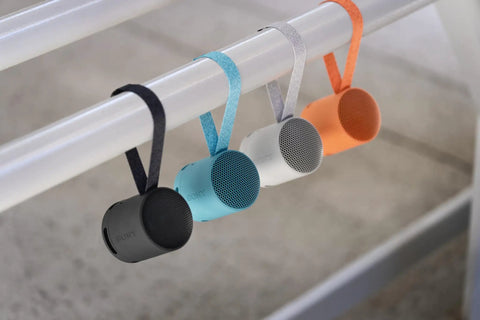 Four XB100 Wireless Bluetooth Portable Speakers in black, blue, gray, and orange with straps hanging on a rod