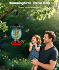 Man and child admiring Solar Powered Hummingbird Feeder With 49 Fl Oz Capacity Hand Blown Blue Glass Color Changing Night Glow Seven Feeding Ports With Perch Never Leaks Outdoor Hanging For Your Yard in a garden setting.