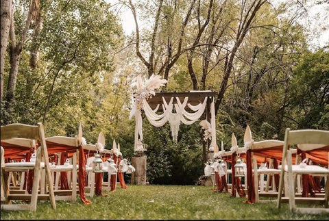 Outdoor wedding aisle with chairs and arch in a wooded setting, decorated with white drapes