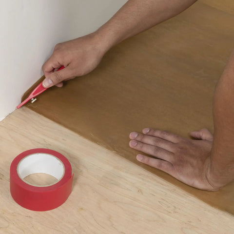 Trimming waxed paper underlayment with red knife during wood floor installation; Waxed Paper Underlayment For Wood Flooring 3 Ft By 250 Ft Roll 750 Sq Ft 30 Lb Pro Grade Heavy Duty Kraft Paper Moisture Barrier To Ease Nail Down Installations And Minimize Squeaks visible.