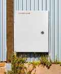 Rugged Outdoor Electrical Box With Lock And Mounting Plate Heavy Duty Steel Junction Enclosure 12 By 10 By 6 Inches NEMA 4X IP66 Waterproof Dustproof Weatherproof Project Box Built To Last For Tough Outdoor Installations installed on a corrugated metal wall with plants below.