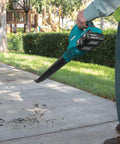User holding Badass 36V Cordless Leaf Blower Kit with two 18V batteries clearing debris off sidewalk in a park.