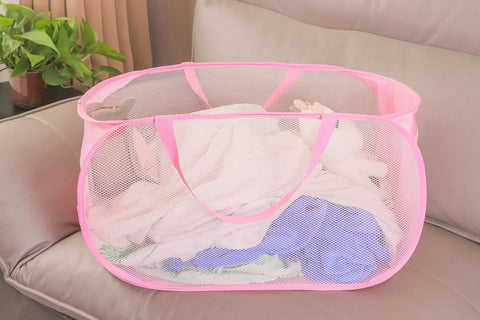 Pink foldable mesh laundry basket pop up hamper with reinforced handles on a sofa, filled with clothes for travel or laundry room.