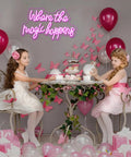 Two girls at a tea party under 'Where The Magic Happens' pink neon sign LED word light for wall decor USB powered, enhancing ambiance.
