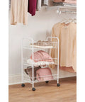 Three Tier Wheeled Wire Shelving Cart With Adjustable Metal Shelves in a bedroom, holding clothes and bags, white finish.
