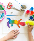 Child using yellow glue on rainbow craft, colorful materials and scissors in view