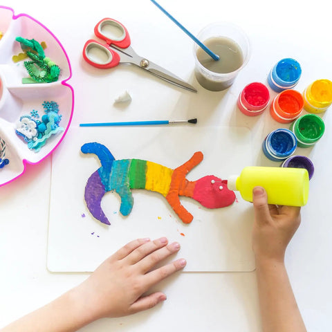 Child using yellow glue on rainbow craft, colorful materials and scissors in view
