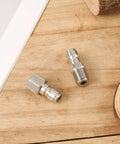 Two stainless steel 1/4-inch quick-connect pressure washer fittings with threaded ends on wooden surface