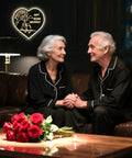Elderly couple in matching pajamas celebrating anniversary with heart-shaped sign and red roses on table in cozy living room.
