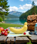 Reusable Banana Keeper Guard With Secure Snap Lid Keeps Bananas Fresh And Prevents Bruising For Tailgate Days Camping Trips And Backyard Hangouts on picnic table with fruits and backpack.