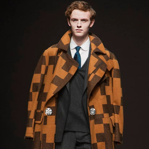 Model wearing a brown and black checkered coat with large lapels, over a white shirt and tie, fastened with snap buttons.