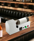 Electric PVC Wine Bottle Capsule Heat Shrinker on a bar top, sealing a wine bottle with a white sleeve for home brewing.