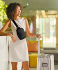 Woman in white dress with Sling Bag Crossbody Fanny Pack For Men And Women With Switchable Strap at airport, suitcase beside her.