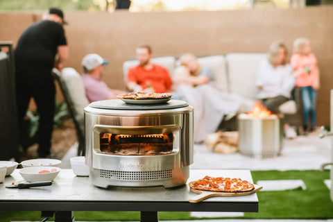 Pizza Oven Starter Bundle With Stainless Steel Outdoor Pizza Maker And Wood Fired Assembly on backyard table, pizza on peel.