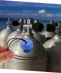 Hand holding blue dive tank valve accessory scuba valve protector cap beside tanks on the beach, emphasizing durability and easy install.