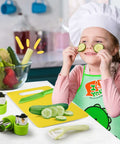 Toddler kitchen tool set with green utensils on yellow cutting board; child in chef hat and apron holds cucumber slices.