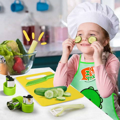 Toddler kitchen tool set with green utensils on yellow cutting board; child in chef hat and apron holds cucumber slices.