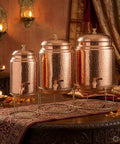Three copper water dispensers with hammered finish and joint free body, brass stands and taps, 5 liter capacity, displayed on table