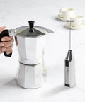 Compact Stovetop Espresso Maker And Handheld Electric Milk Frother Gift Set Pro Grade Aluminum Moka Pot with battery frother in hand.
