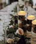Pack Of 4 Wax Flickering LED Floating Candles 3 Inch Tealights Waterproof Warm White Battery Operated Flameless Candles For Dope Centerpieces Weddings Pool Parties Decor in glass vase with pinecones on an elegant table setting.