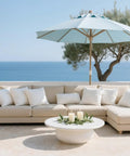Outdoor patio setting with beige couch, white pillows, and a blue umbrella overlooking the sea.