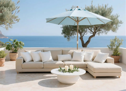 Outdoor patio setting with beige couch, white pillows, and a blue umbrella overlooking the sea.