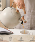 Person pouring tea from a Stove Top 2.5 Quart Whistling Tea Kettle With Food Grade 18 8 Stainless Steel Wood Pattern Folding Handle Creamy White Finish Fast Heating Easy Clean Includes Silicone Heat Insulation Pad into a mug