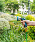 High Pressure Metal Garden Hose Nozzle With 10 Spray Patterns And Thumb Control On Off Valve For Car Wash Plants Pets And Outdoor Cleaning Built For Home Use And Heavy Duty Tasks Blue being held and used to spray water on garden plants.