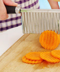 Stainless steel crinkle cutter with black handle slicing orange sweet potato on a wooden cutting board.