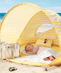 Baby sleeping in Pop Up Baby Beach Tent With 2026 Upgraded UV Protection, yellow stripes, on sandy beach with bag and toys nearby.