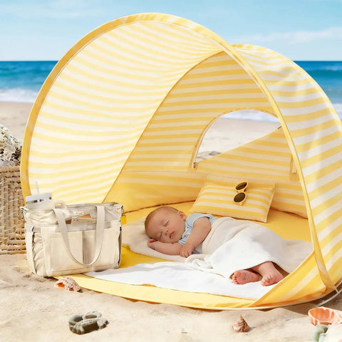 Baby sleeping in Pop Up Baby Beach Tent With 2026 Upgraded UV Protection, yellow stripes, on sandy beach with bag and toys nearby.
