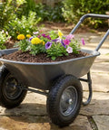 Heavy Duty Garden Wheelbarrow Cart With Dual Wheels And Built In Stand hauling soil and colorful flowers on a stone path