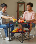 Two people sitting at Folding Camping Table For Outdoor Adventures Portable Lightweight Picnic Table With 66 Lb Capacity Waterproof 600D Oxford Fabric Four Cup Holders Carry Bag Tailgating Camping Beach Trips with snacks and drinks