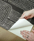 Hands peeling back Nikki Chu Black And White Zulu Signature Peel And Stick Wallpaper Roll, showcasing bold modern pattern.