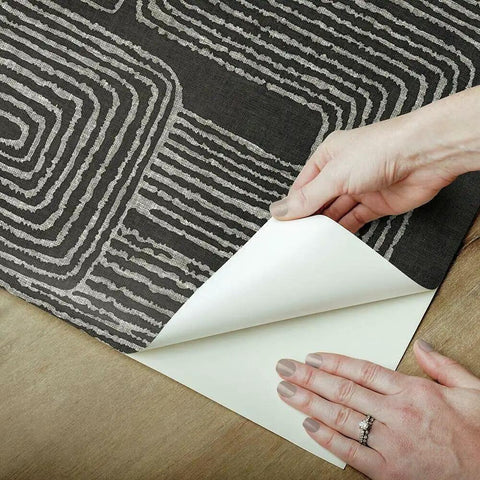 Hands peeling back Nikki Chu Black And White Zulu Signature Peel And Stick Wallpaper Roll, showcasing bold modern pattern.
