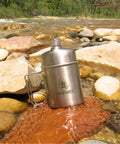 Stainless Steel Nesting Cup And M34 Scout Canteen Set For Camping And Hiking resting on river rocks