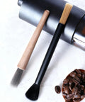 Two brushes from the Dual Head Coffee Grinder And Espresso Machine Cleaning Brush With Stiff Bristles For Deep Ground Residue And Grinder Gunk And Soft Bristles For Gentle Dusting On Surfaces set shown alongside a coffee grinder and beans.